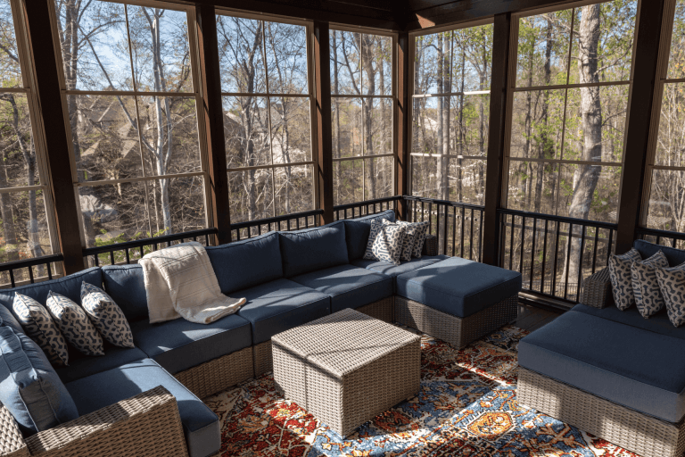 A modern screened-in porch with floor-to-ceiling windows and black metal railings. The space features a large L-shaped sectional sofa with navy blue cushions and wicker frame. The sofa is adorned with decorative pillows in gray and white geometric patterns. A large beige wicker ottoman serves as a coffee table in the center of the seating area. The floor is covered with a large area rug featuring intricate patterns in red, blue, and beige tones. The rug has traditional design elements with floral motifs. Through the windows, there is a view of bare winter trees and a wooded area. Natural sunlight streams through the windows, creating dappled shadows on the furniture and floor. The ceiling is dark-colored, and the overall color scheme consists of navy blue, beige, and natural wood tones. The space is well-lit with both natural and ambient lighting, creating a cozy indoor-outdoor living environment.