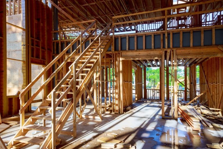 A construction site interior showing a wooden-framed building under construction. A diagonal wooden staircase with handrails is positioned on the left side of the frame. The structure features exposed wooden beams and support posts creating a grid-like pattern throughout. Natural sunlight streams in from above, casting dramatic shadows on the wooden floor and creating a pattern of light and dark areas. The floor is unfinished with scattered construction debris and materials. The ceiling shows exposed rafters and trusses. The walls are partially framed with rough-cut lumber. Through a window opening in the background, green foliage is visible outside. The space has a high ceiling height typical of residential construction. The wooden framing creates a warm, golden tone throughout the image, contrasting with the cool shadows. The construction is in the early stages, with basic structural elements in place but no finished surfaces or fixtures.
